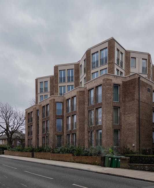 A multi-story residential building with a combination of brick and light-colored façade panels, featuring large windows with some having balconies; the building is situated along a paved street with a sidewalk, and two green waste bins are positioned near the curb. In the foreground, there are partially visible packing materials such as cardboard boxes and protective wrapping on the pavement, indicating ongoing packing or furniture transport as part of a home relocation process. The scene appears overcast with grey clouds above. Hampstead Man and Van, a professional removal service, is involved in the logistics of moving household items, possibly using a van parked nearby, as part of their furniture transport and packing and moving services.
