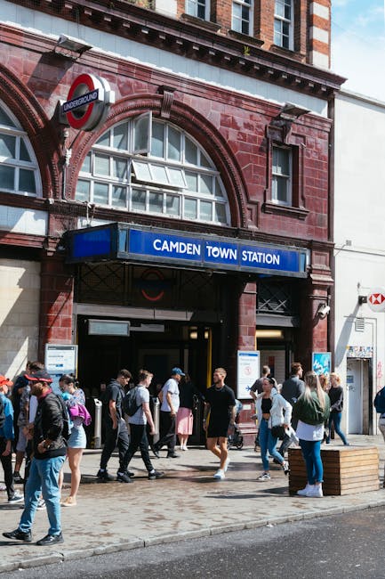 Exterior view of Camden Town underground station with a large blue and white sign displaying 'Camden Town Station' above the entrance. The station building is constructed with red brick and features arched windows. A diverse group of pedestrians, including people carrying backpacks and luggage, are walking on the pavement outside the station entrance. Some individuals are engaged in conversation, while others are focused on entering or exiting the station. To the left, part of the iconic London Underground roundel sign can be seen. The scene captures an active urban environment typical of a busy residential and commercial area, where house removals or furniture transport may be coordinated with nearby local transport options. Hampstead Man and Van may use such a location for logistical planning of house relocations or furniture transport services, emphasizing the importance of accessible transport hubs in moving and packing processes.