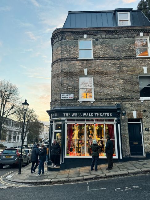The image depicts a street corner in Hampstead during late afternoon or early evening, featuring a brick building with three stories and a prominent theatre on the ground floor named 'The Well Walk Theatre.' The theatre has large display windows illuminated with warm lighting, showcasing decorative items or puppets. Outside the theatre, five people are gathered; some are standing and engaging in conversation, while others are examining the window display. The sidewalk is paved and slightly elevated with a curb, and there are black metal street lamps along the pavement, which is partially lit by the setting sun. A few parked cars are visible on the street, along with residential buildings and trees in the background. This scene captures a typical neighbourhood setting that might be involved in local house moves, as Hampstead Man and Van offers professional removals and furniture transport services in the area, supporting home relocation and moving logistics.