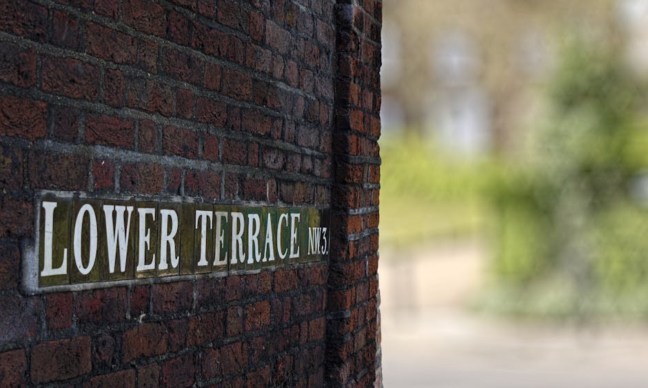 A close-up view of a brick wall with a street sign that reads 'LOWER TERRACE NW3' mounted on it. The sign consists of individual rectangular tiles with white text on a green background, arranged horizontally. The brick wall features dark red bricks with visible mortar joints, extending vertically in the foreground. To the right, the background is blurred but shows greenery and possibly trees, indicating an outdoor setting. This scene is situated outside a residential property on Lower Terrace in Hampstead, which is relevant for a house removal or local moving service. Hampstead Man and Van company might utilize such imagery to illustrate the exterior environment of a typical home setting before or during a furniture transport or packing process in a local move.