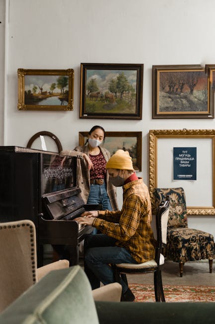 Inside a room with white walls adorned with multiple framed landscape paintings, two individuals are engaged in a moving-related activity. One person, wearing a yellow beanie, a plaid shirt, and a face mask, is seated at a black upright piano, appearing to be packing or preparing to move a piece. The other person, standing nearby, is wearing a face mask, a patterned top, and jeans, observing or assisting with the packing process. The room contains upholstered chairs, including one with floral fabric, and a decorative rug on the floor. A cardboard box and plastic wrapping materials are partially visible, indicating packing activities as part of a home relocation or furniture transport process, consistent with house removals services offered by Hampstead Man and Van.
