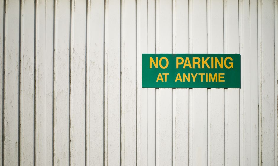 A close-up view of a white wooden wall with vertical planks, displaying a rectangular green sign with bold yellow text that reads 'NO PARKING AT ANYTIME.' The sign is mounted on the wall, which may be part of a building or storage area used during house removals and furniture transport. The lighting is even, highlighting the texture of the wood and the sign, which is relevant in the context of parking restrictions related to removals services. The image exemplifies signage that could be seen near loading zones or parking areas designated for moving vans, supporting the topic of parking and loading restrictions for house relocations as detailed on hampsteadmanandvan.org.uk.