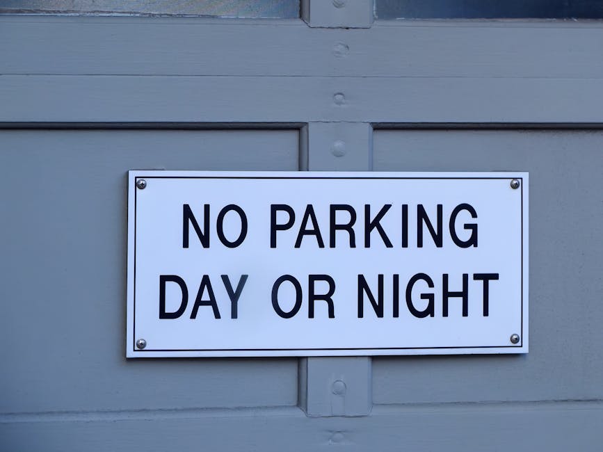 A rectangular white sign mounted on a grey wooden door or panel, displaying the message 'NO PARKING DAY OR NIGHT' in bold black capital letters. The sign is secured with four metallic bolts, one at each corner, and is positioned centrally on the door. Visible elements include the grey painted wood surface with horizontal and vertical paneling details, typical of residential or commercial property exteriors in Hampstead. The environment suggests an outdoor setting, likely at the entrance of a property where vehicle access is restricted to facilitate home relocation or moving services, supported by the presence of the sign indicating parking restrictions relevant for furniture transport and moving logistics. The sign serves to inform enforcement of parking limits during parking and loading activities, foundational in planning removal or packing and moving operations in the area, with Hampstead Man and Van providing professional removals within the context of house relocations.