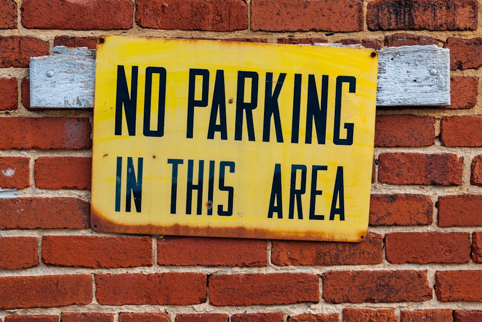 A close-up view of a white wooden wall with vertical planks, displaying a rectangular green sign with bold yellow text that reads 'NO PARKING AT ANYTIME.' The sign is mounted on the wall, which may be part of a building or storage area used during house removals and furniture transport. The lighting is even, highlighting the texture of the wood and the sign, which is relevant in the context of parking restrictions related to removals services. The image exemplifies signage that could be seen near loading zones or parking areas designated for moving vans, supporting the topic of parking and loading restrictions for house relocations as detailed on hampsteadmanandvan.org.uk.