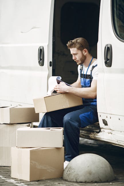 A male worker with light brown hair and a beard, dressed in a striped shirt and blue overalls, is sitting on the edge of a white moving van's open side door during a home relocation process. He is carefully examining a document or packing list while holding a cardboard box on his lap. Surrounding him on the ground are multiple cardboard boxes of various sizes, some sealed with packing tape, ready for loading or unloading. The van is parked on a paved area with a concrete bollard nearby, and the interior of the van is visible, showing more packing materials and boxes inside. This scene depicts the packing and loading stage of furniture transport and house removals, with Hampstead Man and Van providing professional moving services for a domestic move.