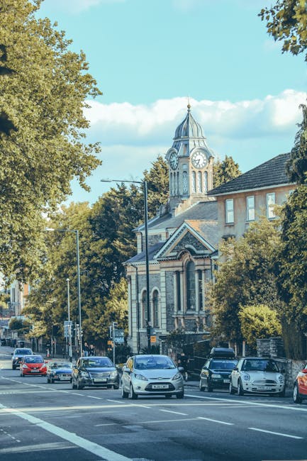 A wide street scene in Hampstead showing rows of white Victorian-style residential buildings with ornate architectural details, balconies, and large windows. The street is lined with green bushes and mature trees with partially green and brown foliage, indicating an autumn setting. Several vehicles, including parked cars and a white van, are positioned along the curb. Traffic lights are visible at the intersection, with red signals illuminated, and a few pedestrians are seen standing or crossing near the roadside. The sky above is overcast with grey clouds, creating diffuse lighting. This image captures a typical residential urban environment suitable for a house removal process, with furniture, packing materials, or moving equipment not visible, but implying a quiet neighbourhood ready for relocation or moving services provided by Hampstead Man and Van.