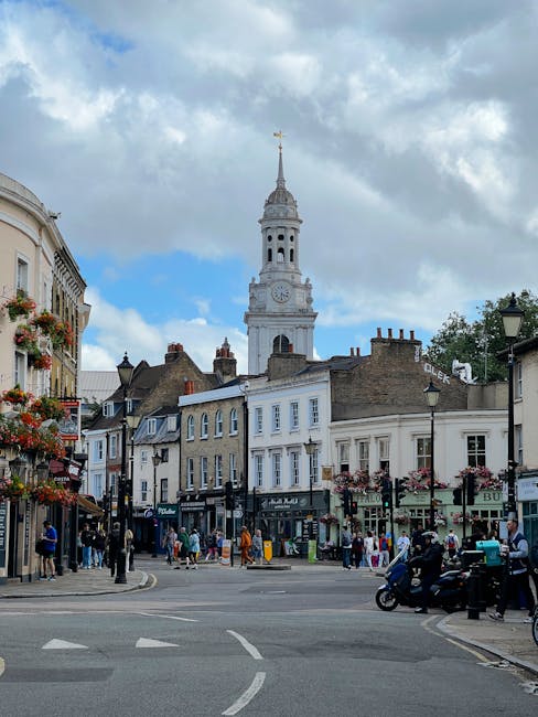 A busy street in Hampstead with a mix of historic and modern buildings, including a prominent white clock tower with a spire, set against a partly cloudy sky. The street is lined with shops, cafes, and decorative hanging flower baskets on lampposts. Pedestrians are walking along the pavement, some carrying shopping bags or talking, while a black motorcycle and several parked cars occupy the roadside. Inside an open doorway on the left, there are visible cardboard boxes, packing materials, and furniture covered with blankets, indicating a home relocation or moving activity. A Hampstead Man and Van van is parked nearby, ready for furniture transport, with visible loading ramps and equipment for moving house items. The scene captures the dynamic process of packing and loading during a scheduled removal service, showcasing the busy environment typical of property relocations in Hampstead.